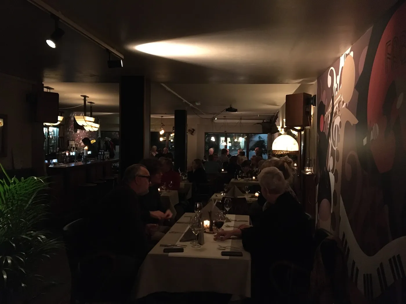 Ingetogen verlicht dinergedeelte bij De Fabel met de jazz-muurschildering op de achtergrond